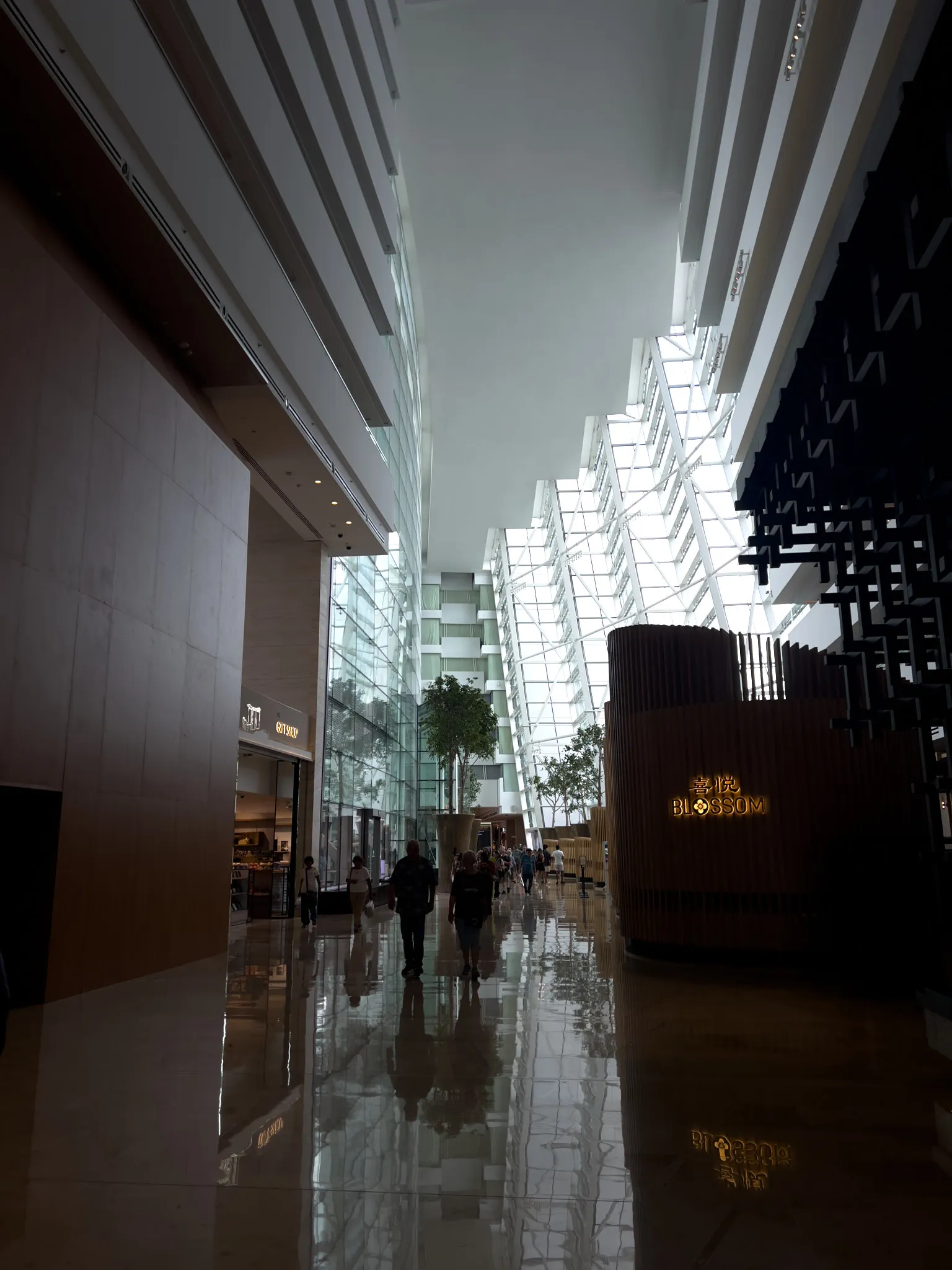 A spacious, modern atrium with tall glass walls allowing abundant natural light to flood the interior. The high ceiling is lined with white horizontal architectural beams, and the polished floor reflects the surroundings and people walking through the space. Indoor trees in large planters add greenery along the central walkway. Shops and restaurants line the sides, including a warmly lit sign that reads 'BLOSSOM' on the right. The overall atmosphere is open, airy, and elegant.