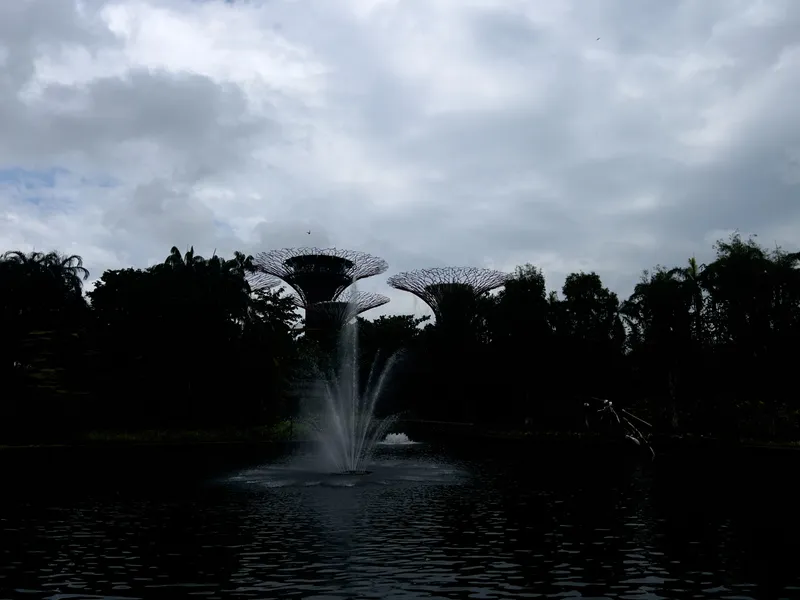 A dark, reflective pond sits in the foreground with a fountain spraying water upward from its center. Behind the pond, dense vegetation forms a shadowed tree line. Rising above the trees are tall, sculptural tower-like structures with open lattice tops. The sky is filled with thick, overcast clouds, creating a dramatic contrast against the silhouetted landscape.
