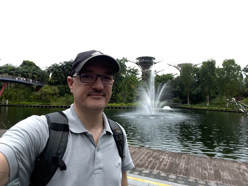 A person takes a selfie while standing on a wooden boardwalk beside a calm body of water with a large fountain spraying upward in the background. Behind the fountain, dense tropical greenery and tall tree-like structures rise above the landscape. A curved pedestrian bridge with several people walking on it appears to the left, while trees and thick foliage frame the scene. The person wears a light gray polo shirt, a dark cap, glasses, and a backpack.