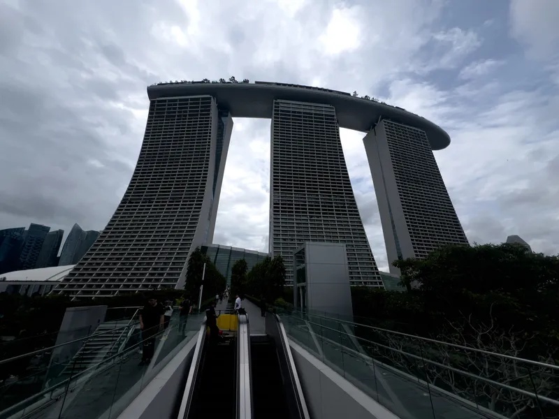 Three tall, curved towers connected by a long rooftop structure stretch upward beneath a cloudy sky. A glass-lined pedestrian walkway and escalator lead toward the base of the buildings, with a few people walking along the path. Lush greenery borders the walkway, and additional modern skyscrapers are visible in the distant background.