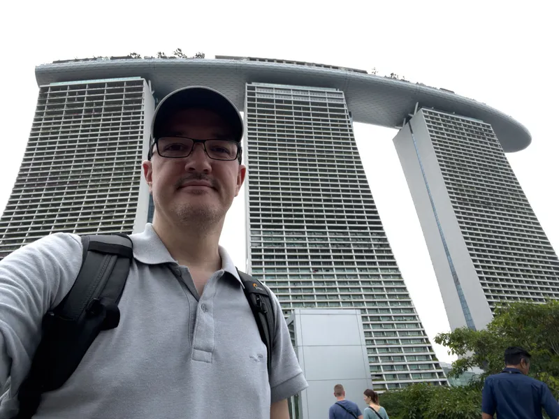 Man wearing a light gray polo shirt, glasses, and a backpack standing outdoors with three tall, modern high‑rise towers behind him, connected at the top by a long, curved rooftop structure. Several other people are visible in the background near greenery, and the sky appears overcast.