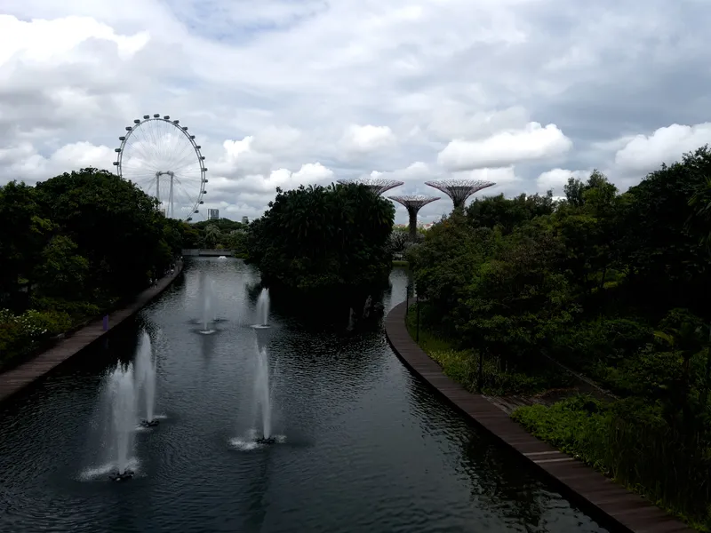 A long, calm waterway lined with multiple fountains stretches between dense, green foliage on both sides. Wooden walking paths run along the edges of the water, curving gently through the lush landscape. In the distance, a large observation wheel rises above the treetops, while several towering tree‑shaped structures stand to the right. Soft, overcast clouds fill the sky, casting diffused light over the scene.