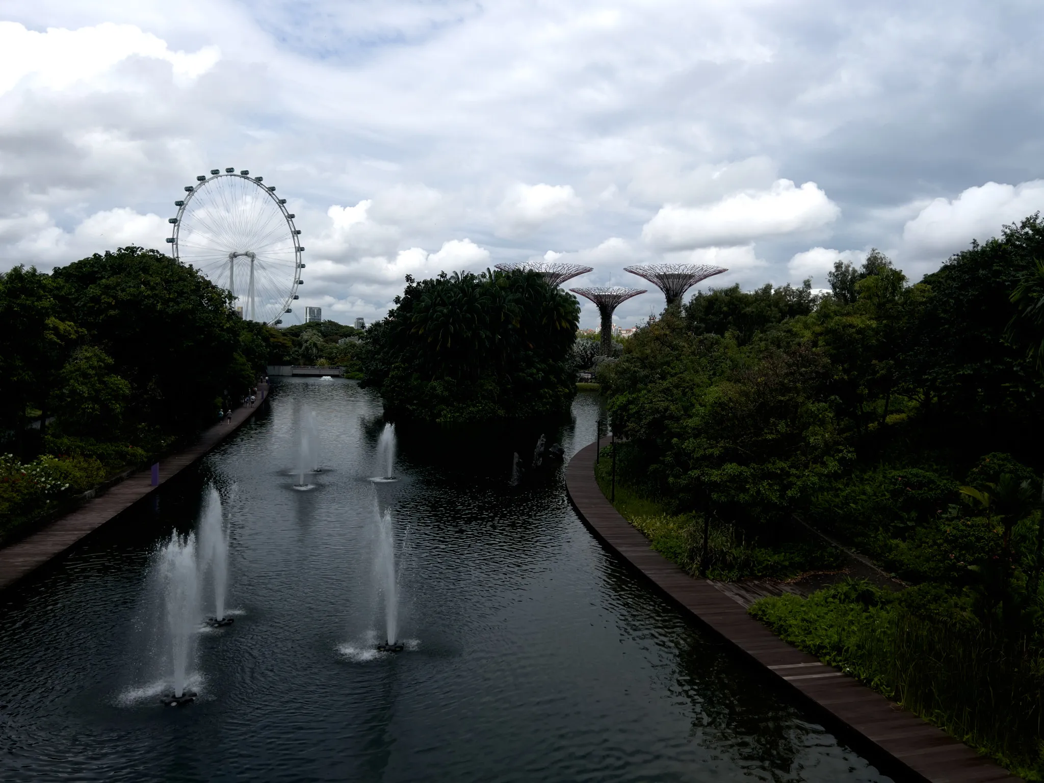 A long, calm waterway lined with multiple fountains stretches between dense, green foliage on both sides. Wooden walking paths run along the edges of the water, curving gently through the lush landscape. In the distance, a large observation wheel rises above the treetops, while several towering tree‑shaped structures stand to the right. Soft, overcast clouds fill the sky, casting diffused light over the scene.