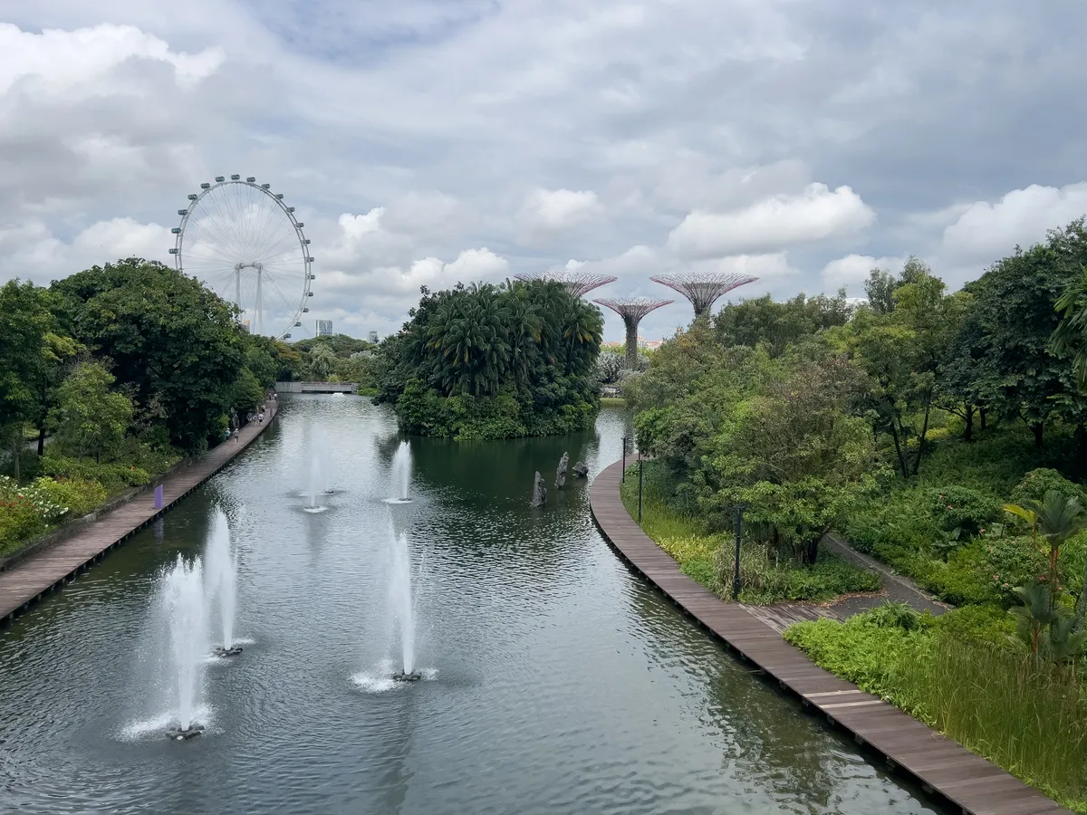 In the Civic District, Downtown Core, Central Region, Singapore, a picturesque scene stretches along a calm waterway. Fountains bubble up from the centre, surrounded by lush greenery and varied vegetation. To the left in the background looms Singapore's large Ferris wheel, while to the right the striking, futuristic Supertree structures rise into the sky. A lightly clouded sky gives the scene a peaceful atmosphere.