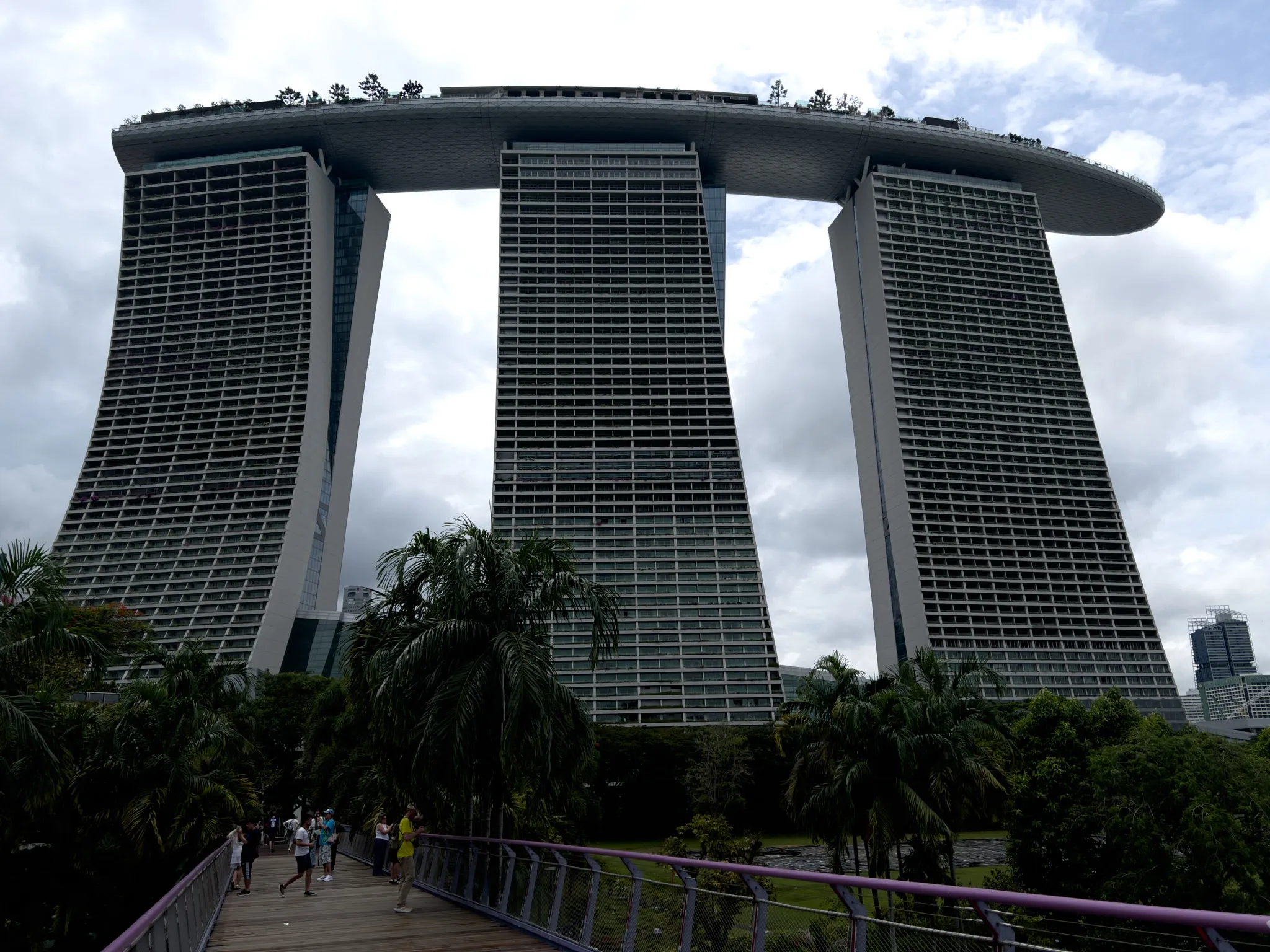 Three tall, curved high-rise towers connected by a large rooftop structure with trees and greenery stretch upward under a cloudy sky. A pedestrian walkway lined with purple railings leads through lush tropical plants and palm trees toward the towers. Several people walk along the path, adding a sense of scale to the massive architectural complex.