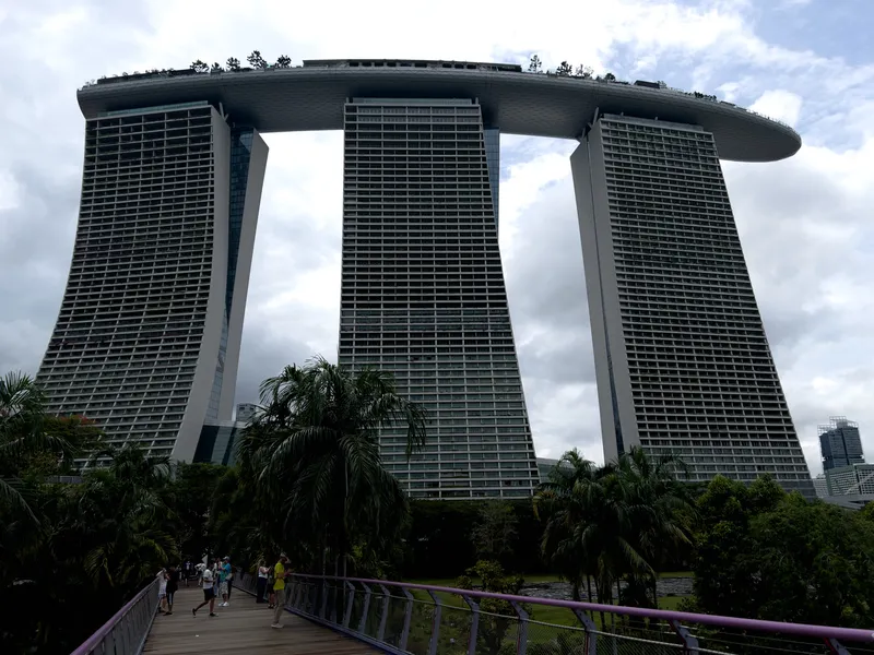 Three tall, curved high-rise towers connected by a large rooftop structure with trees and greenery stretch upward under a cloudy sky. A pedestrian walkway lined with purple railings leads through lush tropical plants and palm trees toward the towers. Several people walk along the path, adding a sense of scale to the massive architectural complex.