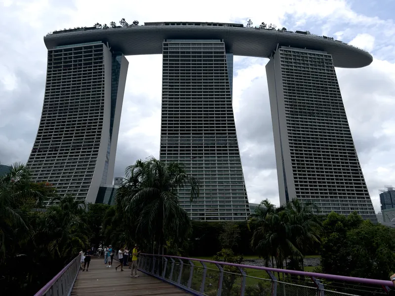 Three curved high‑rise towers connected at the top by a long, boat‑shaped SkyPark, rising above a landscaped area with tall palms and a wooden pedestrian walkway where several people are walking. The scene is framed by overcast skies and dense greenery in the foreground.