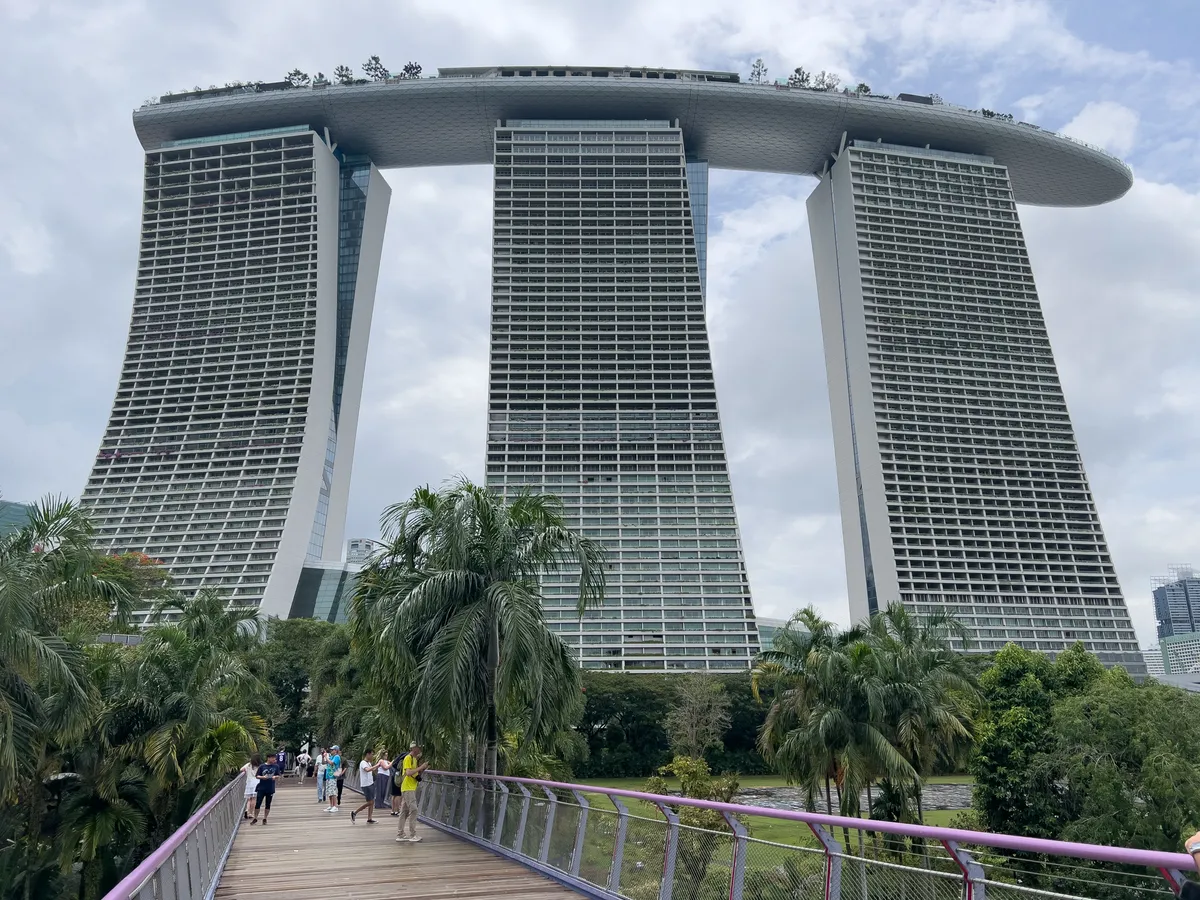 The image captures the impressive architecture of Marina Bay Sands in the Civic District, Downtown Core, Central Region, Singapore. Its three striking towers stand asymmetrically and are linked by a large, suspended platform on which small trees and plants grow. In the foreground, a wooden bridge carries pedestrians past lush palms and green spaces that enrich the cityscape. A lightly clouded sky makes the modern structure of the buildings stand out all the more.