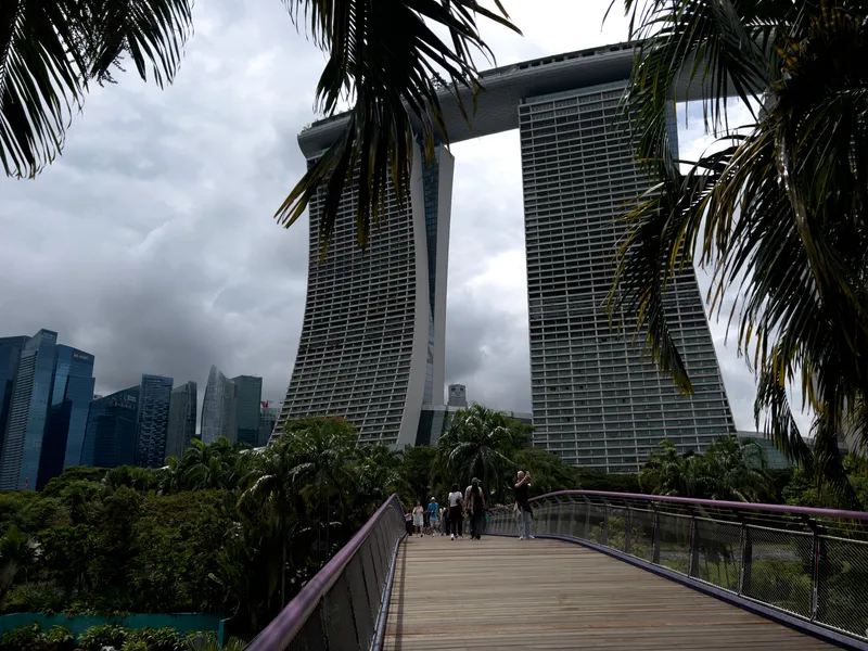 A wooden pedestrian walkway leads through lush tropical greenery toward three tall, curved towers connected by an elevated skybridge. Palm leaves frame the scene from above, and several people walk along the path. Dark, heavy clouds loom in the sky, contrasting with the modern city skyline in the distance.