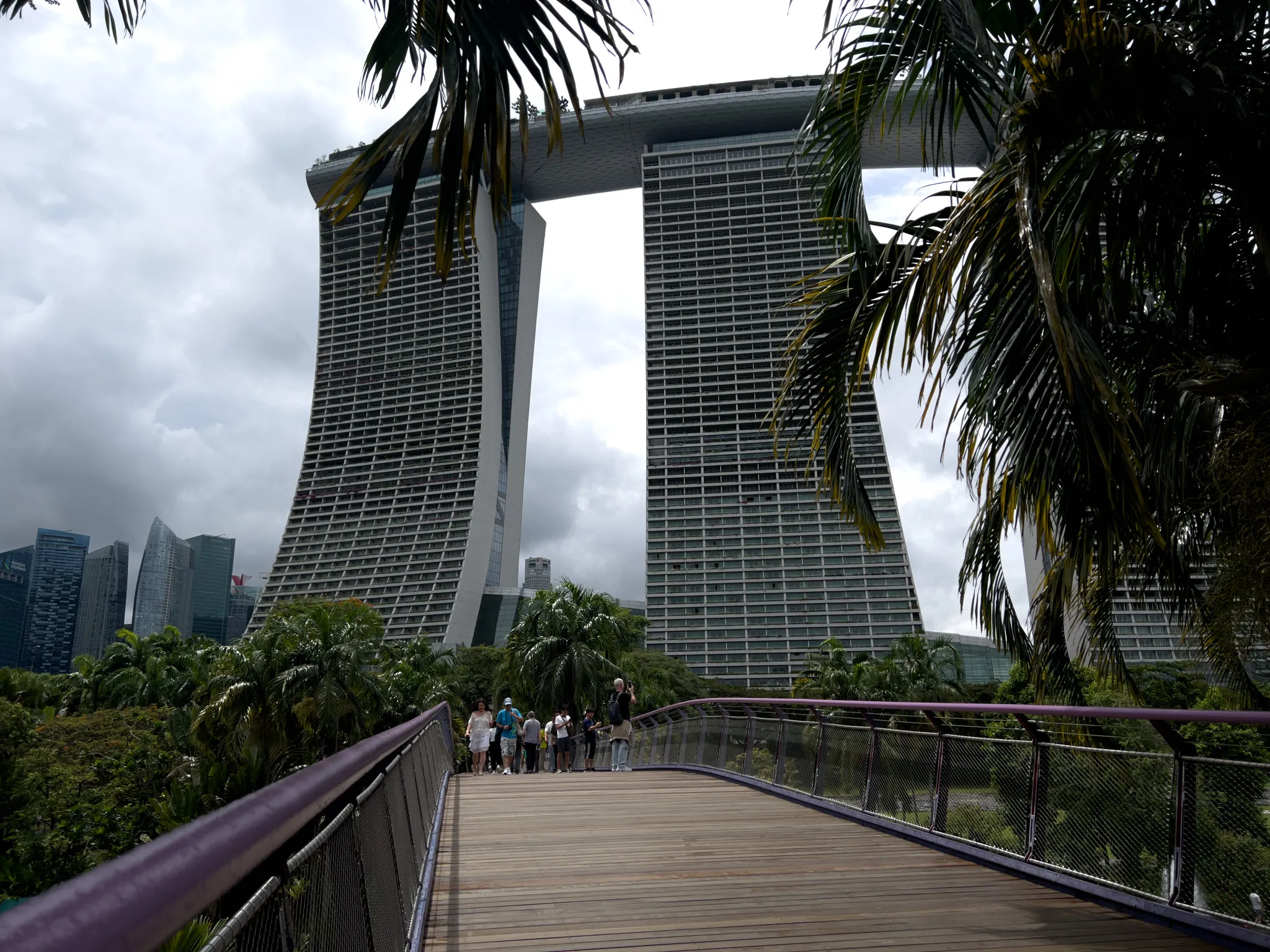 A curved wooden walkway leads toward three tall, interconnected high-rise towers with distinctive outward-leaning shapes and a long skybridge spanning across their rooftops. Dense tropical plants and palm trees frame the scene, with a group of people gathered at the end of the walkway. The sky is overcast, casting diffused light over the modern cityscape and surrounding greenery.