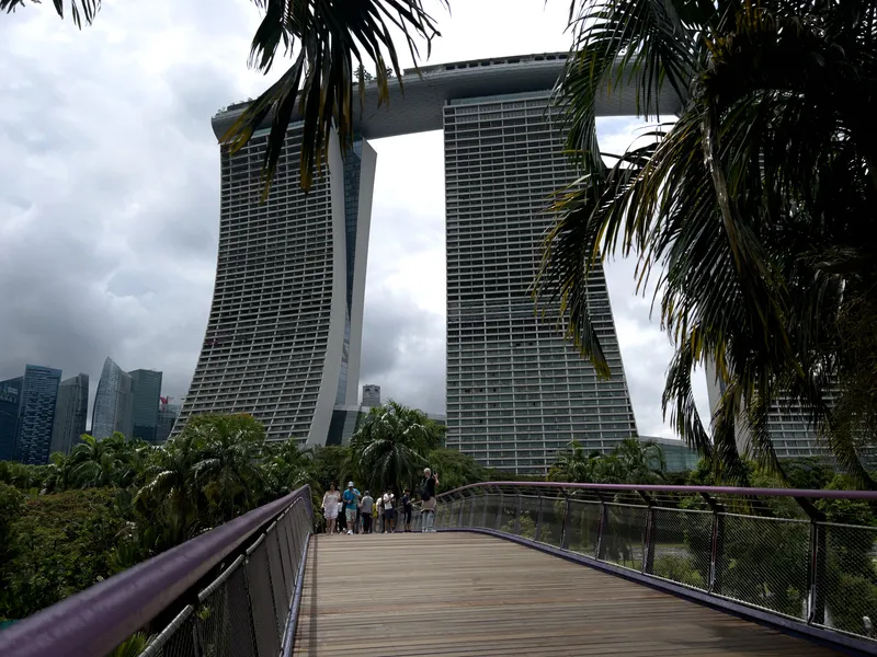 A curved wooden walkway leads toward three tall, interconnected high-rise towers with distinctive outward-leaning shapes and a long skybridge spanning across their rooftops. Dense tropical plants and palm trees frame the scene, with a group of people gathered at the end of the walkway. The sky is overcast, casting diffused light over the modern cityscape and surrounding greenery.