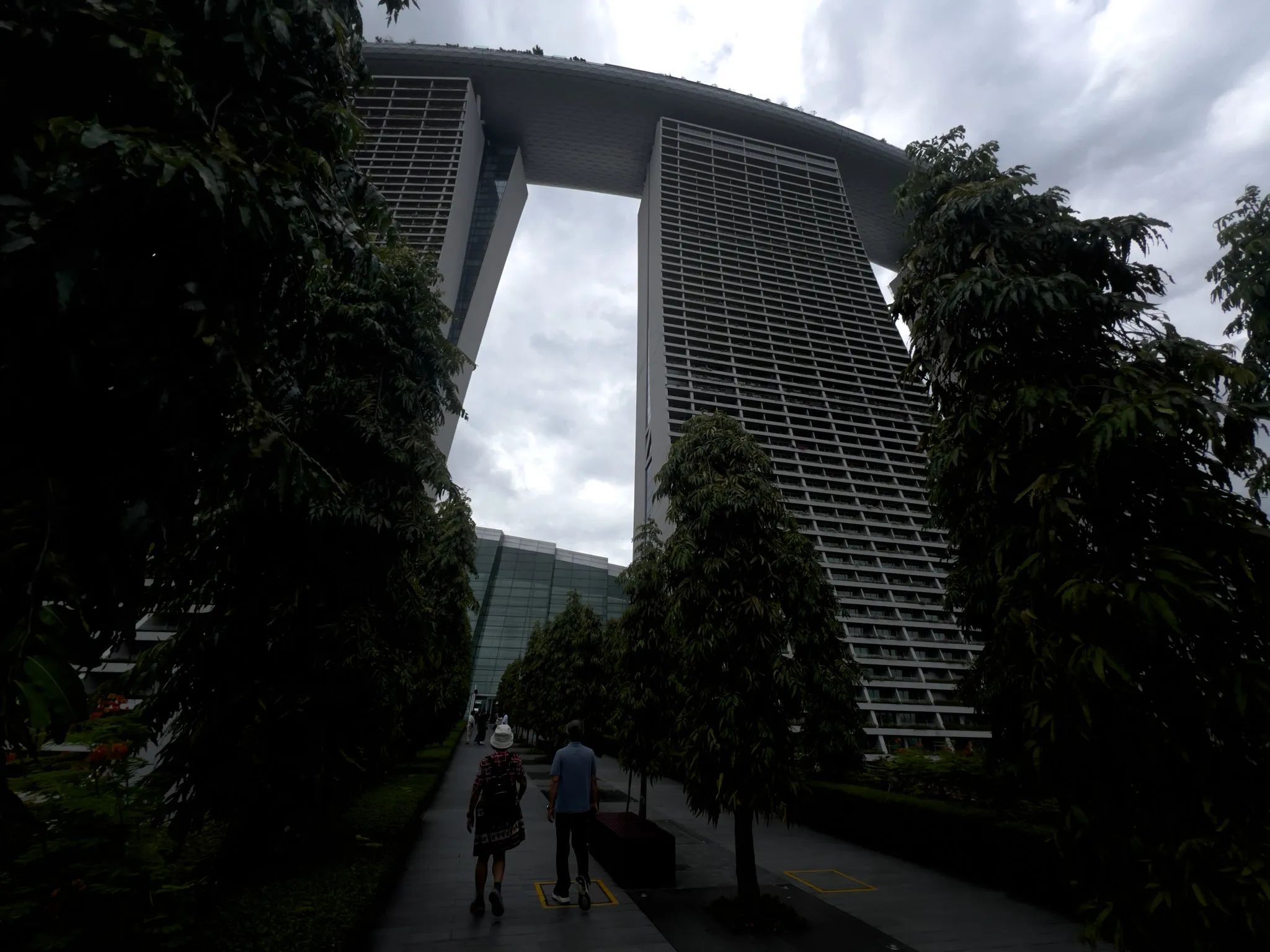 A tree-lined walkway leads toward a towering three-part high-rise structure connected at the top by a large, curved rooftop deck. Two people walk along the path, surrounded by tall, dense greenery, while the massive building rises above them under an overcast sky.