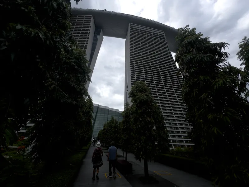 A tree-lined walkway leads toward a towering three-part high-rise structure connected at the top by a large, curved rooftop deck. Two people walk along the path, surrounded by tall, dense greenery, while the massive building rises above them under an overcast sky.