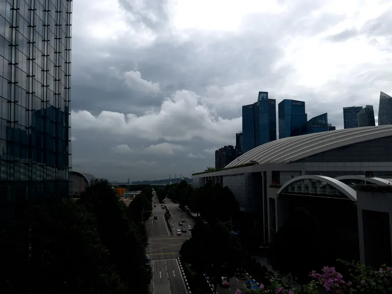 A wide urban scene featuring a long, tree-lined road running through the city toward the horizon. The sky is filled with dark, heavy clouds suggesting an incoming storm. On the left stands a tall glass building reflecting the cloudy sky, while on the right are modern curved-roof structures and several blue high-rise towers. Vehicles travel along the multi-lane road, and patches of greenery and flowers appear in the foreground.