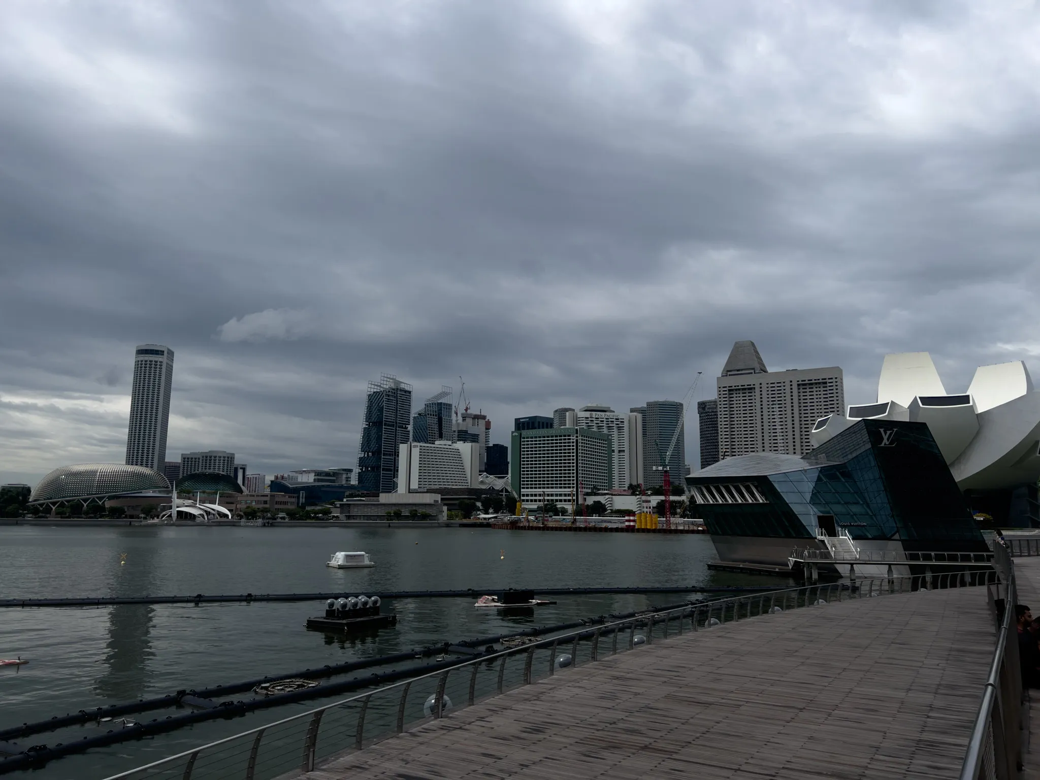 Curved wooden boardwalk running alongside calm bay water, facing a dense cluster of modern high‑rise buildings under a dark, overcast sky. Distinctive contemporary structures with geometric and glass designs stand near the water’s edge, while additional towers and rounded architectural forms rise behind them. A few floating platforms and small watercraft rest on the surface of the bay. The scene appears quiet and moody, with heavy clouds dominating the background.