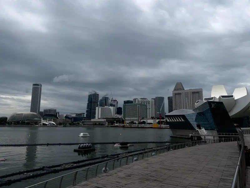 Curved wooden boardwalk running alongside calm bay water, facing a dense cluster of modern high‑rise buildings under a dark, overcast sky. Distinctive contemporary structures with geometric and glass designs stand near the water’s edge, while additional towers and rounded architectural forms rise behind them. A few floating platforms and small watercraft rest on the surface of the bay. The scene appears quiet and moody, with heavy clouds dominating the background.