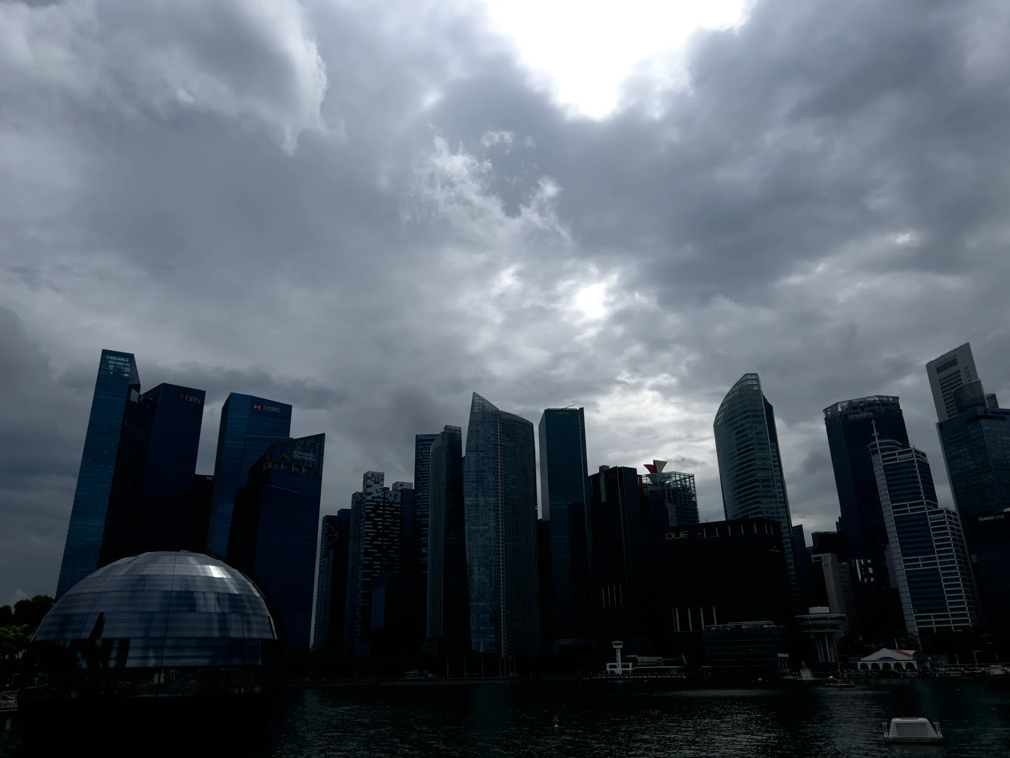 A cluster of tall, modern skyscrapers rises along a waterfront beneath a thick, overcast sky. The buildings form a dark silhouette due to the heavy cloud cover, with only faint reflections and highlights visible on the glass surfaces. In the foreground, a large reflective dome structure sits on the water, mirroring some of the surrounding architecture. The scene feels dramatic and moody, with the bright break in the clouds contrasting sharply against the shadowed cityscape.