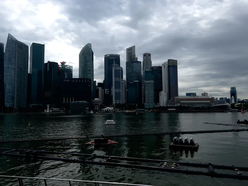 Dense cluster of modern skyscrapers rising along a waterfront under a cloudy, overcast sky, with tall glass buildings forming the main skyline. The water in the foreground is calm, reflecting parts of the darkened structures and floating equipment positioned near the shore. A large building with a red-roofed upper level stands to the right side of the scene, while the overall atmosphere appears muted and shaded due to the heavy cloud cover.