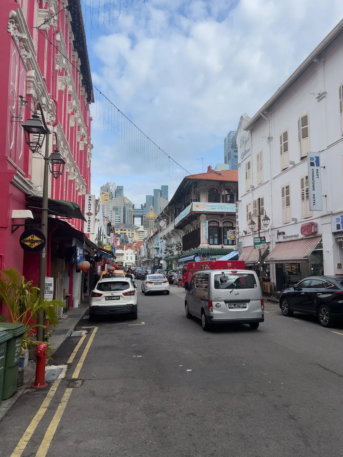 The scene captures a busy street in Tanjong Pagar, Outram, Central Region, Singapore. On the left rises a striking pink building in classical style. The right shows a row of bright, historic facades housing restaurants and shops. The street is lined with parked cars while a few vehicles travel in both directions. In the background, modern skyscrapers contrast with the historic architecture. A lightly clouded sky lends the setting a pleasant atmosphere.