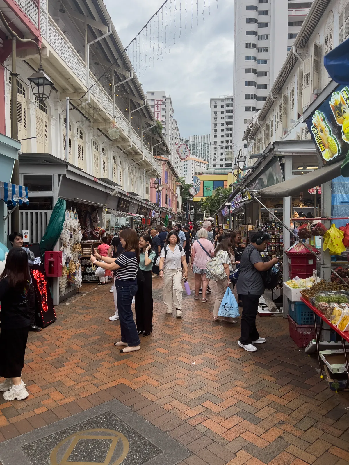 In Tanjong Pagar, Outram, Central Region, Singapore, a busy alley stretches out with colourful historic buildings on both sides. People stroll along the paved street in varied groups, with some pausing at stalls selling souvenirs and snacks. The atmosphere is lively and multicultural, while clouds above the tall buildings in the background add an urban touch.