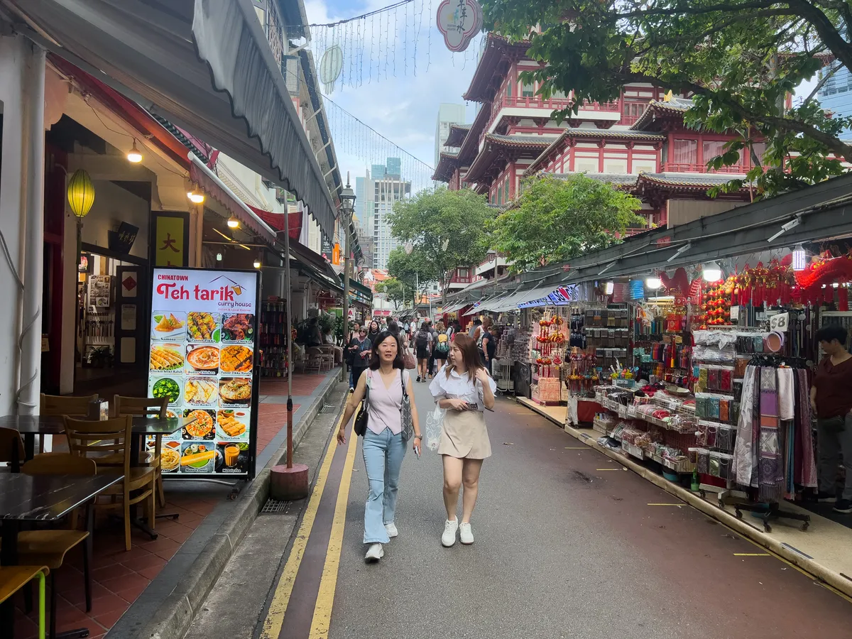 On the busy street of Tanjong Pagar, Outram, Central Region, Singapore, two women stroll at a leisurely pace while numerous stalls and restaurants shape the surroundings. On the left hangs a large poster for a Teh Tarik Curry House showing a selection of traditional dishes. The right of the frame shows colourful stalls packed with souvenirs and decorative items. In the background stands the striking architecture of a red-and-white traditional building, part of Chinatown's iconic look.