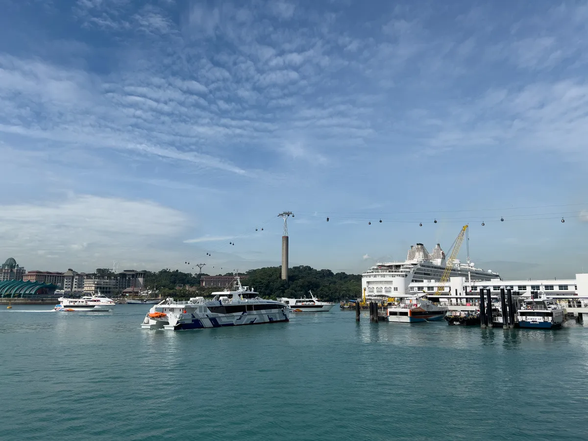 A picturesque view of the harbour at Fort Siloso in the Southern Islands, Central Region, Singapore. In the foreground, several boats rest on the calm, turquoise water. Especially striking is a large white cruise ship moored at a dock. Above the water, a cable car runs with gondolas that swing gently between the hills. In the background, a green, forested island stretches out while the sky is decorated with light white clouds.