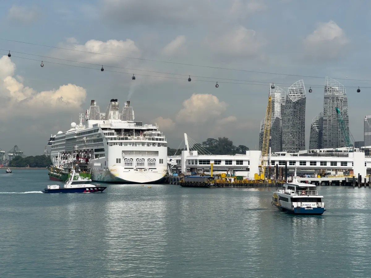 At Fort Siloso in the Central Region of Singapore, a large cruise ship lies moored in the harbour. Beside it, a smaller vessel is heading towards the port. Along the shoreline, several modern skyscrapers stand out with their distinctive forms. Cable cars stretch above the water, while a partly cloudy sky lets gentle sunlight glint off the water.