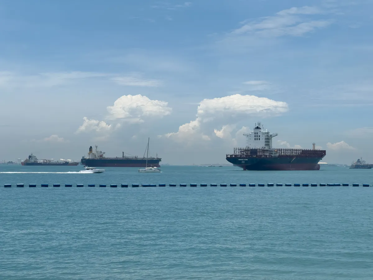 The scene shows several large ships anchored in the water off Fort Siloso in the Southern Islands, Central Region, Singapore. In the foreground, a small boat moves briskly across the calm water while two large cargo ships are clearly visible in the background. The sky is pale blue, with a few white clouds hanging on the horizon. A blue buoy at the edge of the frame marks the boundary between water and shore.