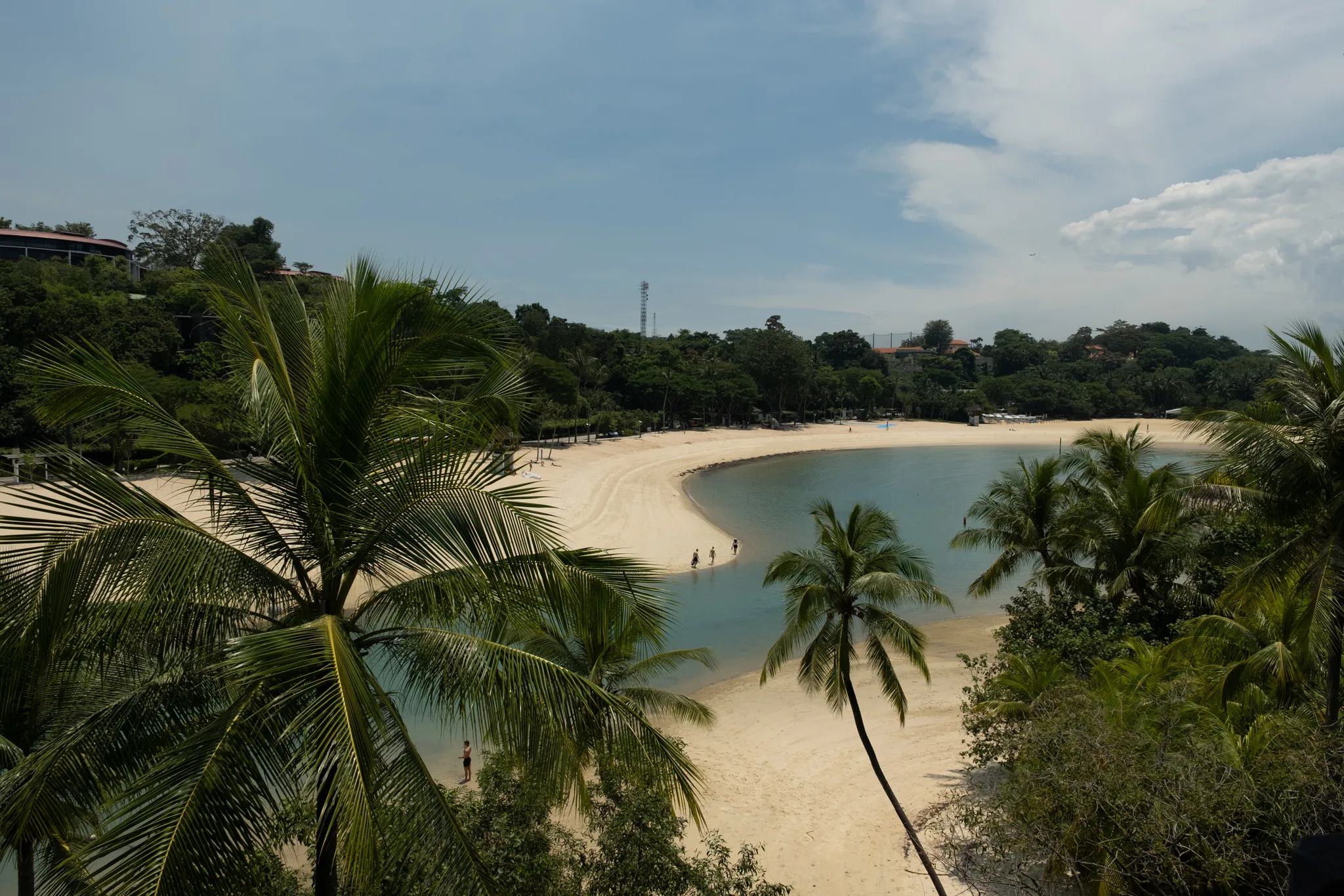 An elevated view of a curved tropical beach with white sand forming a crescent shape around calm, shallow turquoise waters. Tall coconut palm trees frame the foreground and middle ground, their green fronds swaying gently. A few people walk along the water's edge near the sandy shoreline. The beach is bordered by dense tropical vegetation and lush green trees stretching across the background, with some resort-style buildings partially visible among the foliage on both sides. A tall communications tower rises above the treeline in the center background. The sky is mostly blue with scattered white and grey clouds. The scene resembles Palawan Beach or Siloso Beach on Sentosa Island, Singapore.