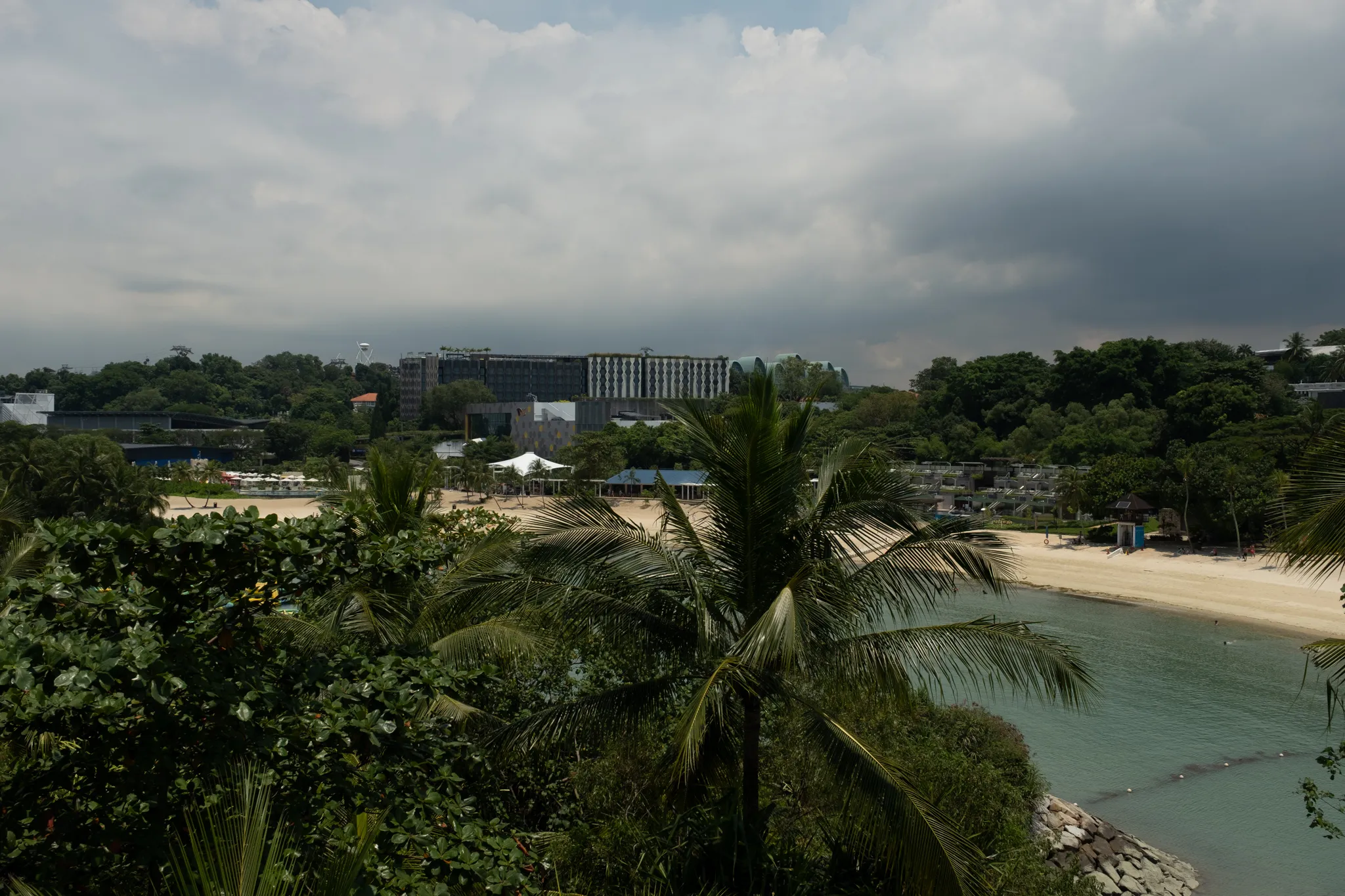 An elevated view of a tropical beachfront on Sentosa Island, Singapore, featuring turquoise waters lapping against a white sandy beach. Lush green palm trees and tropical vegetation dominate the foreground, partially framing the scene. In the middle ground, beach facilities including tented structures and low-rise buildings are visible along the shoreline. Behind them, the distinctive architecture of Resorts World Sentosa, including the Hotel Michael and the Festive Hotel, rises above the tree line. The sky is overcast with dramatic gray and white clouds, suggesting approaching rain or humid tropical weather. The overall scene captures the blend of natural beauty and modern resort development characteristic of Sentosa Island.