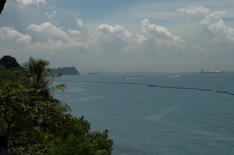 A panoramic coastal view from an elevated vantage point overlooking a calm, blue-green sea. In the foreground, lush tropical vegetation including a tall coconut palm tree and dense green shrubs frame the left side of the scene. A floating barrier or boom line stretches across the water from left to right. Several vessels are visible on the water, including cargo ships and a speedboat creating a white wake. In the background, hazy islands and headlands are partially obscured by atmospheric haze, while large white dome-shaped storage tanks, likely for oil or gas, are visible on the far right shoreline. The sky is filled with towering cumulus clouds against a pale blue backdrop, typical of a tropical Southeast Asian climate. The overall scene suggests a view from Sentosa or the southern coast of Singapore looking out toward the Strait of Singapore and nearby Indonesian islands.