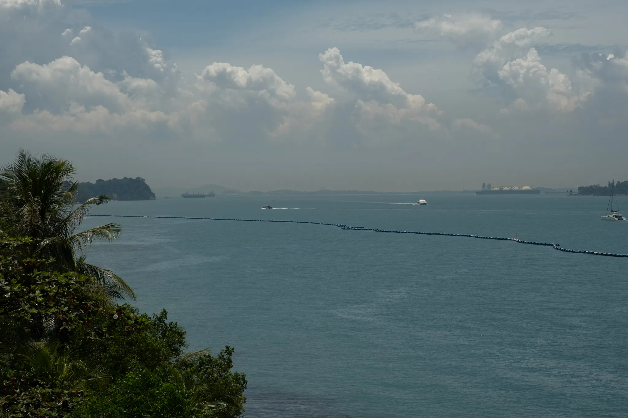 A panoramic coastal view from an elevated hillside looking out over a calm, blue-green sea. In the lower left corner, lush tropical vegetation including a tall coconut palm tree and dense green foliage frames the scene. A long floating barrier or boom line stretches across the water from left to right. Several boats and vessels are visible on the water, including what appears to be a white ferry or speedboat creating a wake. In the hazy distance, small islands and landmasses are visible along the horizon, along with what appear to be industrial storage tanks or port facilities on the far right. Cargo ships can be seen anchored in the background. The sky above is partly cloudy with large white cumulus clouds building up, while a hazy atmospheric layer softens the distant views, suggesting a warm, humid tropical climate.