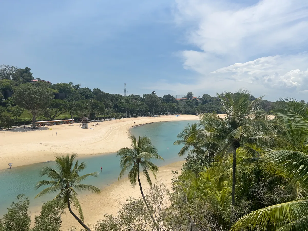 A clear view of a quiet beach at Fort Siloso, Southern Islands, Central Region, Singapore. The white sandy beach with its gentle waves is lined with green palms. In the background, hills rise up with lush vegetation as a few visitors soak up the relaxed atmosphere. A narrow channel of water runs through the beach area, while the partly cloudy sky lets the daylight fall softly on the scene.