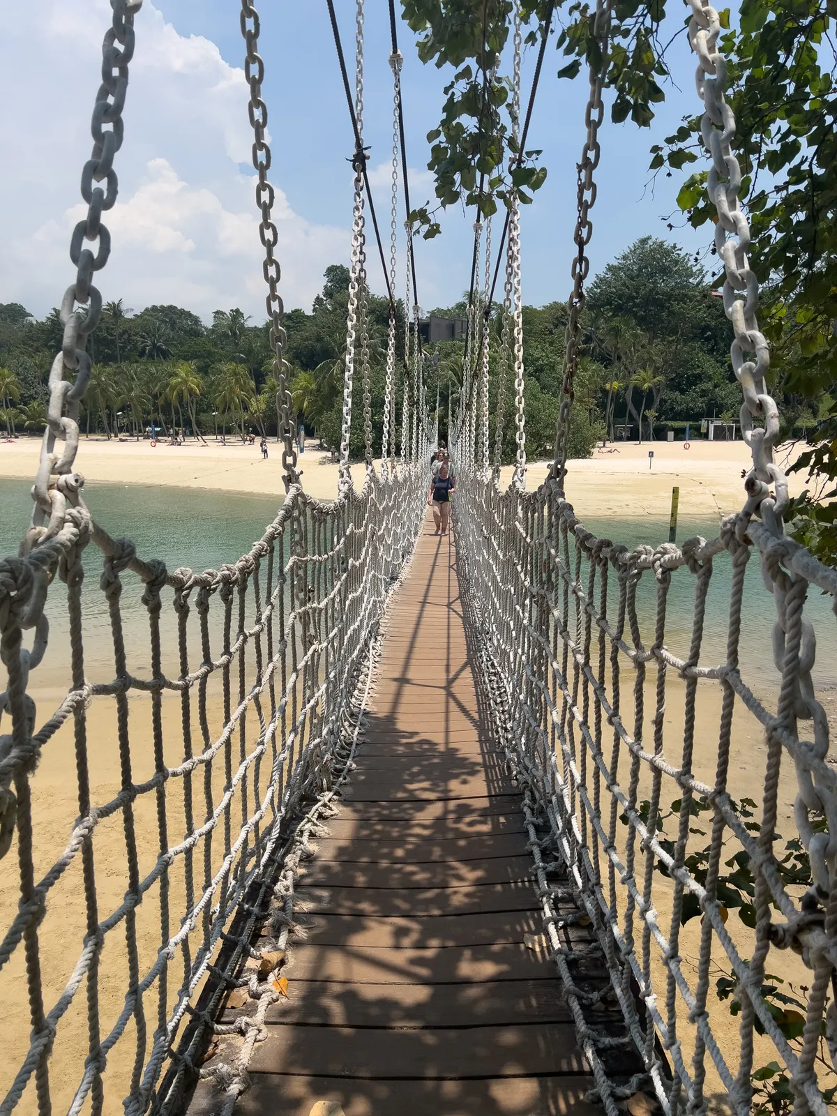 A suspension bridge crosses the water at Fort Siloso, Southern Islands, Central Region, Singapore. Framed by dense green branches, the bridge looks out on a quiet beach lined with palms in the background. The scene is sunny, and the shadows cast by the cables fall across the wooden deck. A single person walks across the bridge towards a bright blue sky dotted with a few clouds.