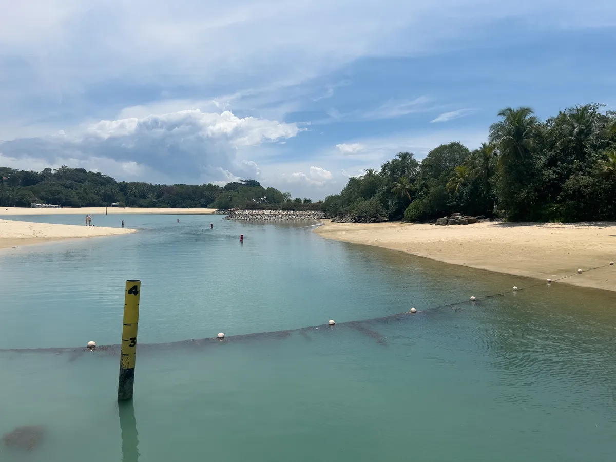 At Fort Siloso in the Southern Islands, Central Region, Singapore, a calm coastal landscape unfolds, with clear turquoise water lapping gently against a sandy beach. Green palms and dense vegetation frame the scene. In the distance, people can be seen enjoying the beach, while a thick, cloudy sky provides a picturesque backdrop. A yellow, numbered bollard stands in the water, swim buoys mark the area and stones enclose the small harbour.