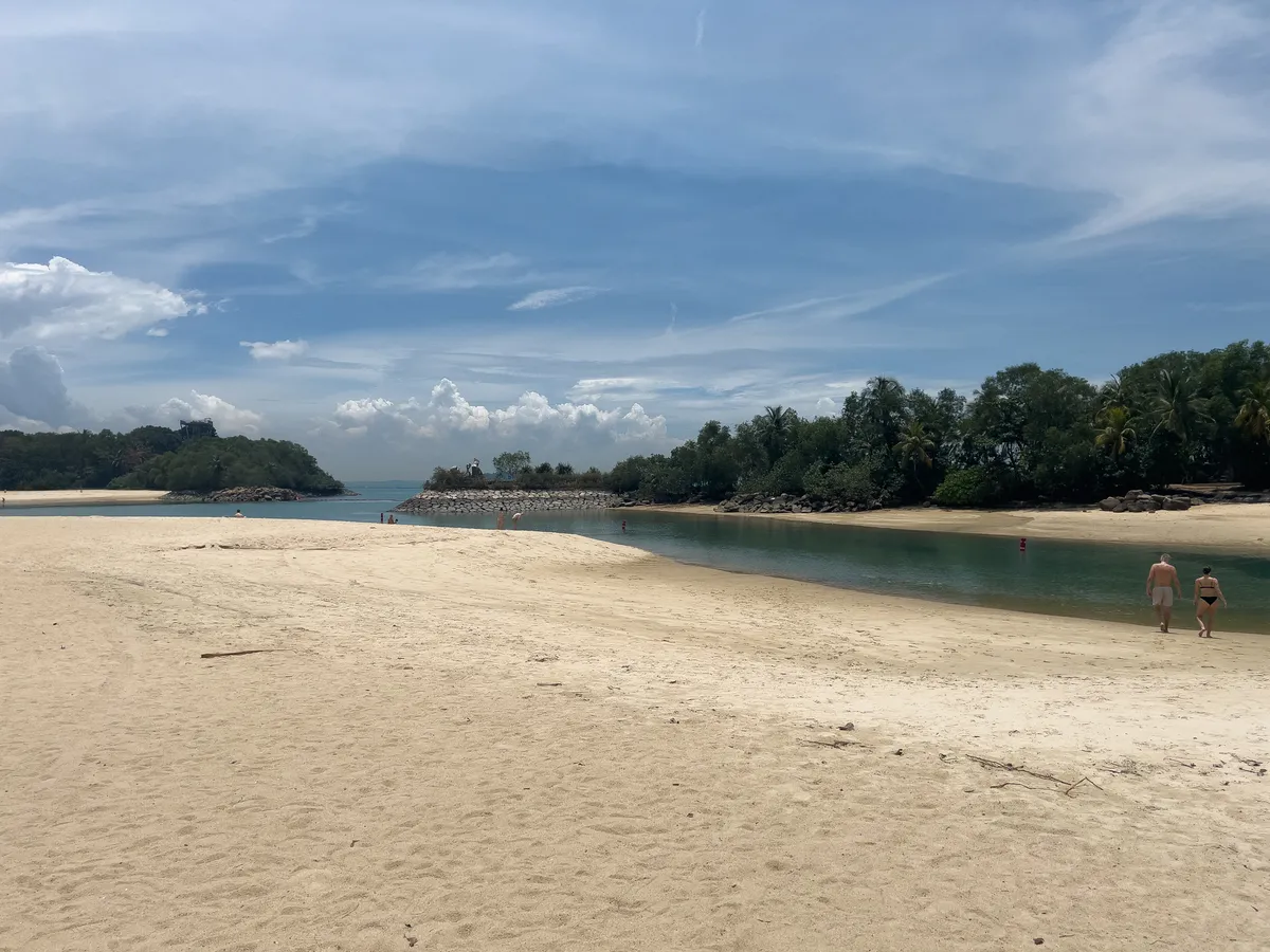 At Fort Siloso beach in Singapore, fine, light sand stretches out beneath a bright blue sky dotted with a few white clouds. Gentle waves roll in on the shore, while a calm lagoon of clear turquoise water shimmers in the background. A wooded coast lines the left of the frame, and on the opposite side a stone wall encloses a small marina. Two people stroll along the shore, taking in the idyllic view. The lush vegetation and shimmering water invite you to spend a relaxing day at the beach. This peaceful scene is located at Fort Siloso, Southern Islands, Central Region, Singapore.