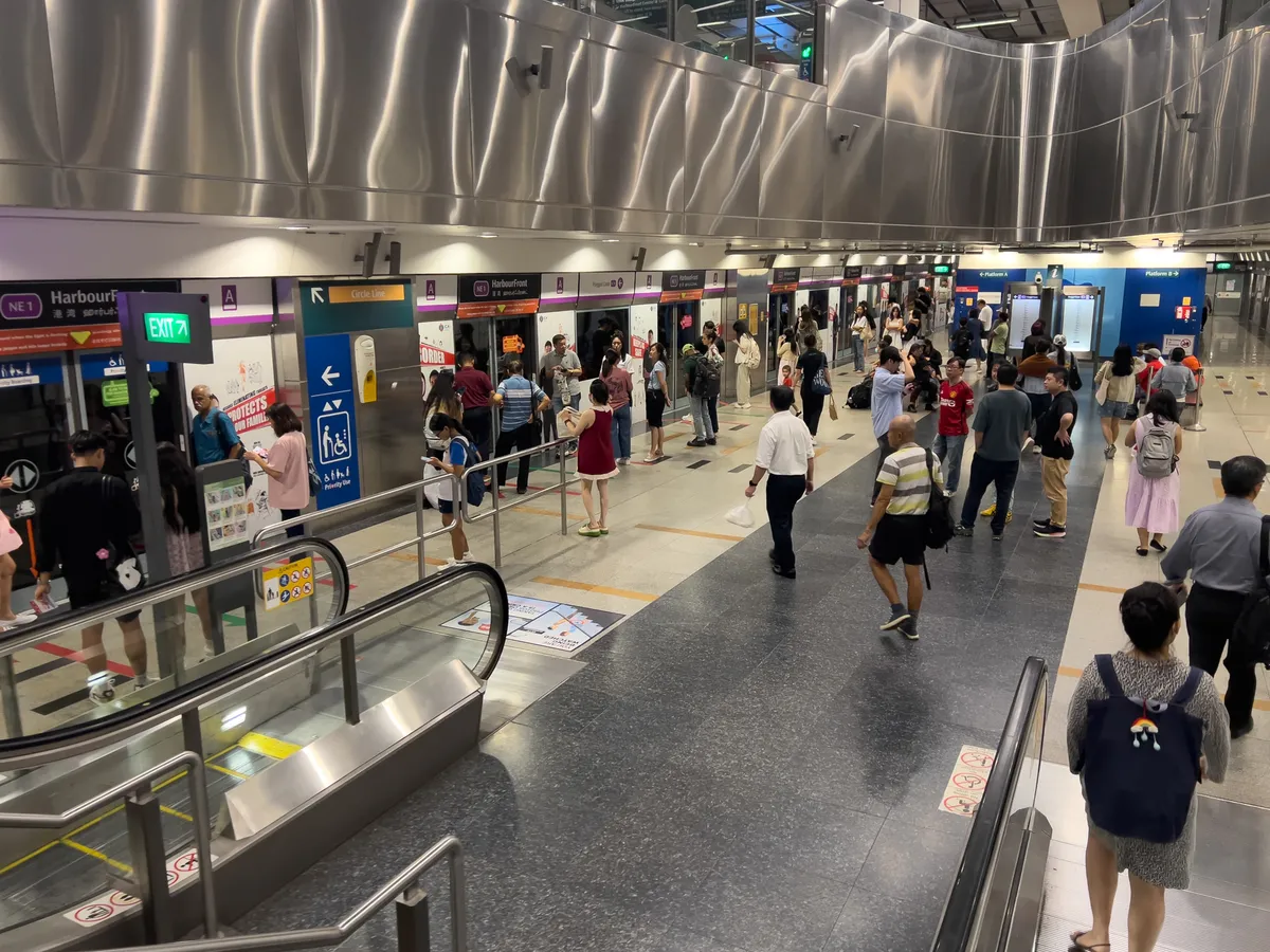 At Berlayar metro station in Bukit Merah, Singapore, a lively scene unfolds. Many people stand on the platforms waiting for their trains while others move through the station. The walls are covered with information and signs about the metro line. On the left, a lift leads up to the upper levels. The interior feels modern, with gleaming metal surfaces and clear, easy-to-read signage. The busy, lively atmosphere reflects the character of urban life in Singapore.
