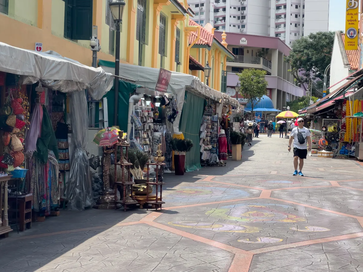 A lively market street in Little India, Rochor, Central Region, Singapore. On the left, several stalls show off colourful textiles and traditional handicrafts. The stalls are decorated with brightly coloured fabrics, inviting you to browse. In the middle of the street, a man in light-coloured clothing and blue shoes strolls past. The walkway is decorated with a colourful, artistic pattern. In the background, tall buildings form a modern contrast to the traditional market atmosphere. Under a white parasol, more passers-by soak up the lively mood.