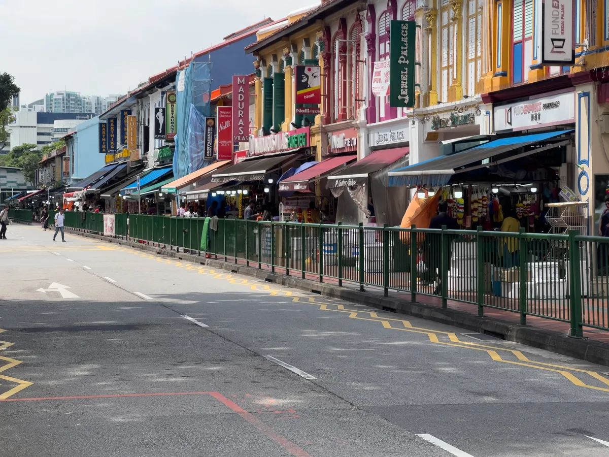 In Little India, Rochor, Central Region, Singapore, a busy shopping street stretches out, lined with colourful, historic buildings featuring wooden shutters and distinctive signage. Numerous shops offer a wide variety of goods while passers-by stroll leisurely along the road. A green metal railing separates the pavement from the carriageway. Above the shops hang parasols in various colours, casting shade. The scene captures the cultural diversity and vitality of a neighbourhood known for its shops and culinary offerings.