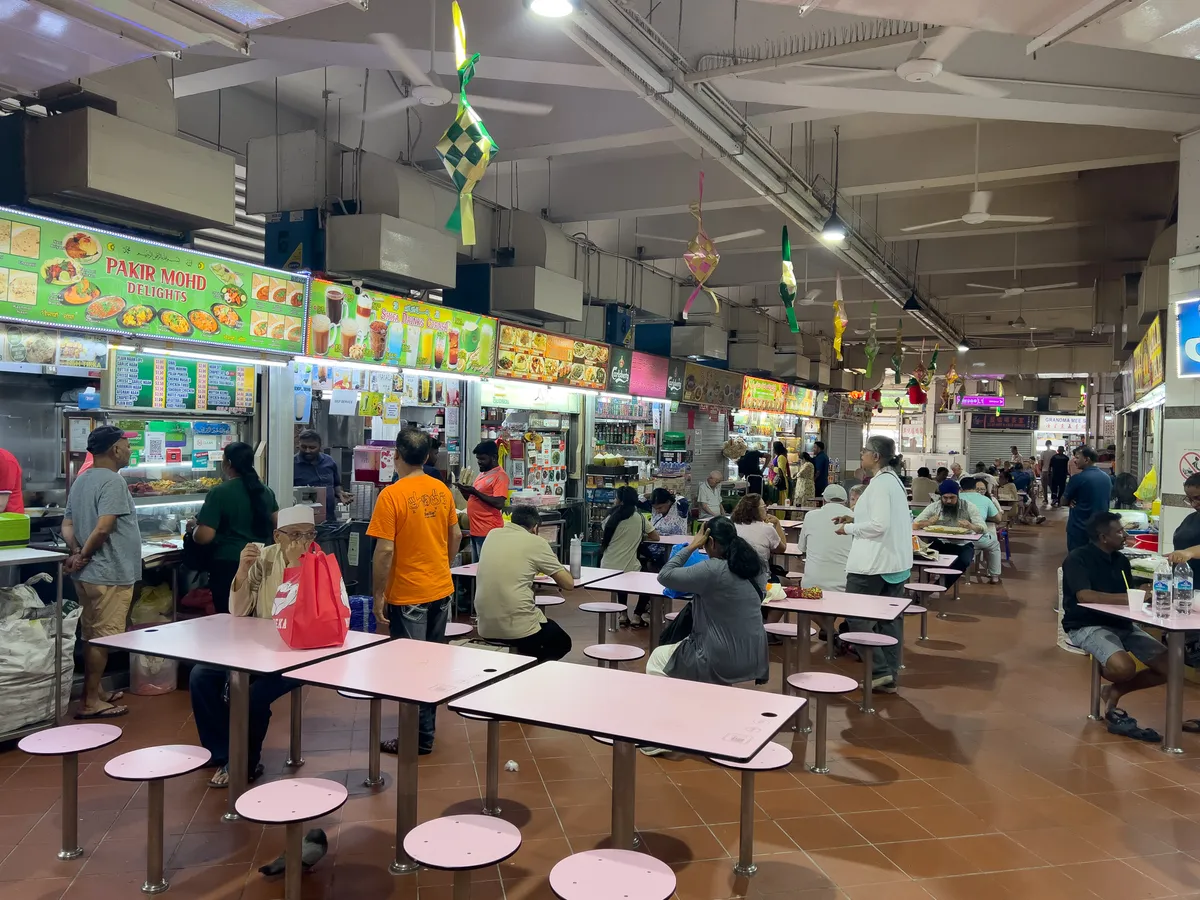 In Little India, 210668, Singapore, the scene shows a busy food hall with colourful stalls offering a range of dishes and drinks. People queue up to choose from an extensive menu while others sit at tables and eat. The tables have pink surfaces, and a few chairs are missing. The lighting is bright, and decorative elements hang from the ceiling. The atmosphere is lively and sociable.