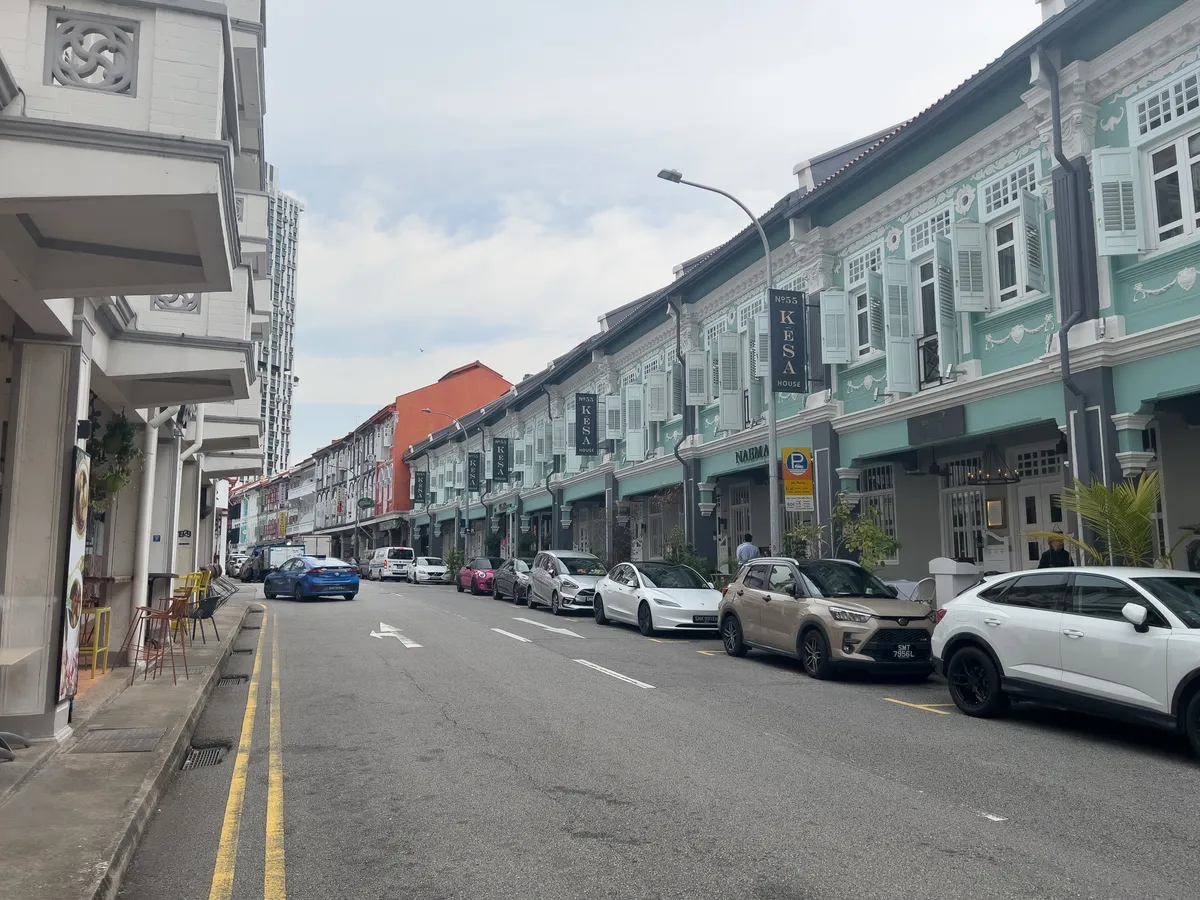 Along a lively street in Tanjong Pagar, Outram, Central Region, Singapore, several historic, colourful buildings with characteristic window shutters are on display. To the left, modern structures underline the area's urban character. The street is lined with cars, including a blue vehicle and several white ones. Small terraced seating areas with chairs and tables create an inviting atmosphere. The buildings show a mix of traditional and contemporary architecture, while the sky above the scene is lightly clouded.