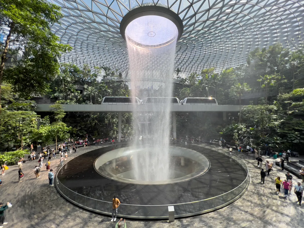 At Jewel Changi in Changi Village, Singapore, a majestic waterfall plunges from the upper dome into a round, level basin. Surrounded by lush green plant life and a unique glass roof structure, a crowd of visitors gathers on the lower platform to take in the spectacular scene. In the background, trains glide across an elevated platform above the space, while bright sunlight streams through the glass roof and fills the room with a warm atmosphere.