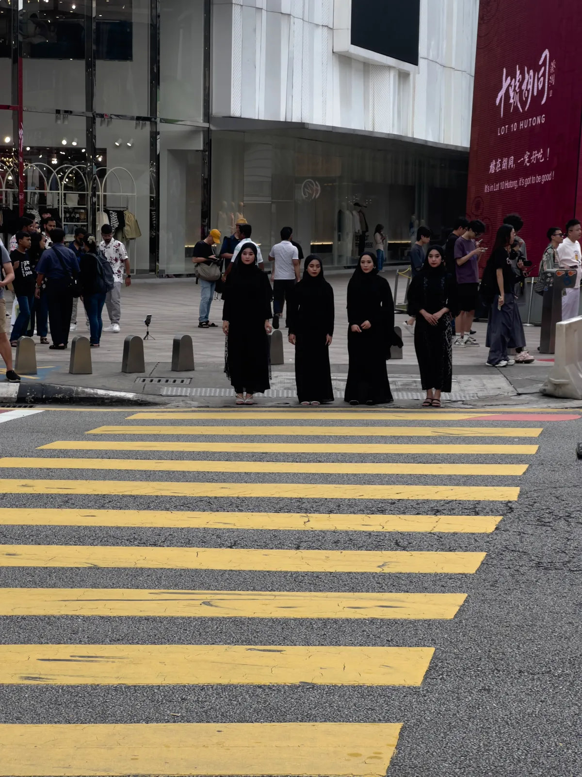 In Kampung Cendana, Kuala Lumpur, four women in black robes stand on a crosswalk. They look into the camera and appear to be coming from a modern shopping area. Numerous passers-by move in different directions in the background, while shop windows display clothing and accessories. The yellow crossing stripes contrast with the grey asphalt, and the scene conveys a sense of urban life and cultural diversity.