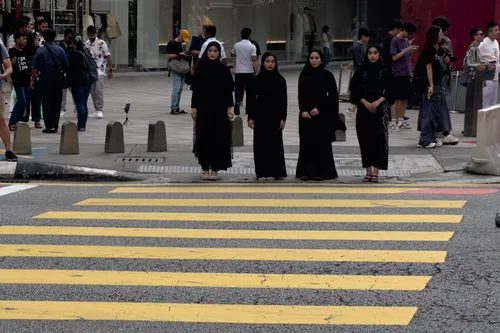 In Kampung Cendana, Kuala Lumpur, four women in black robes stand on a crosswalk. They look into the camera and appear to be coming from a modern shopping area. Numerous passers-by move in different directions in the background, while shop windows display clothing and accessories. The yellow crossing stripes contrast with the grey asphalt, and the scene conveys a sense of urban life and cultural diversity.