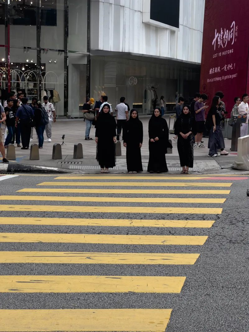 Four young women wearing black hijabs and long black outfits stand together at the edge of a yellow-striped pedestrian crosswalk in Kuala Lumpur's Bukit Bintang shopping district. They appear to be posing for a photo, with a phone on a tripod visible to their left. Behind them, the modern glass-fronted Lot 10 shopping mall is visible, along with a large red banner reading "Lot 10 Hutong" with Chinese characters and the tagline "It's in Lot 10 Hutong, it's got to be good!" Numerous pedestrians of diverse backgrounds walk along the sidewalk in the background. Concrete bollards line the curb separating the road from the walkway. The scene is set on a busy urban street with the characteristic yellow crosswalk markings typical of Malaysian roads.