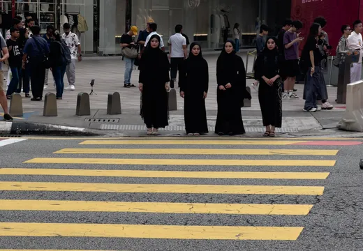 In Kampung Cendana, Kuala Lumpur, stehen vier Frauen in schwarzen Gewändern auf einem Zebrastreifen. Die Frauen blicken in die Kamera und scheinen aus einer modernen Shopping-Umgebung zu kommen. Im Hintergrund sind zahlreiche Passanten zu sehen, die in verschiedenen Richtungen gehen, während Schaufenster mit Kleidung und Accessoires sichtbar sind. Die gelben Zebrastreifen kontrastieren mit dem grauen Asphalt, und die Szene vermittelt ein Gefühl von urbanem Leben und kultureller Vielfalt.