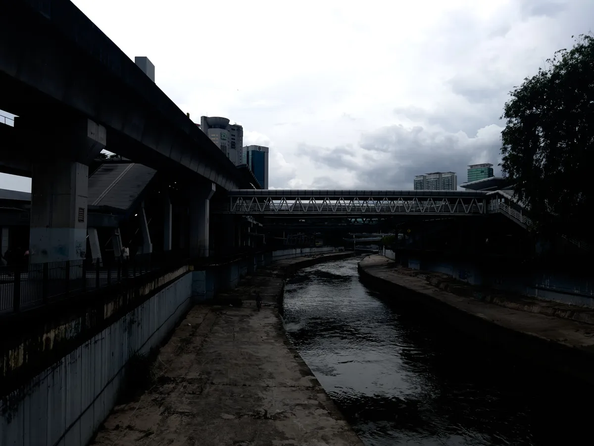 A view of a narrow waterway running through Kampung Cendana in Kuala Lumpur. On the left, massive concrete bridges and modern buildings are visible, partly hidden by dark clouds that create a mystical atmosphere. The bridges seem to float above the water, while uneven concrete surfaces line the banks. Scattered plants grow along the edges, and the entire scene is bathed in muted light.