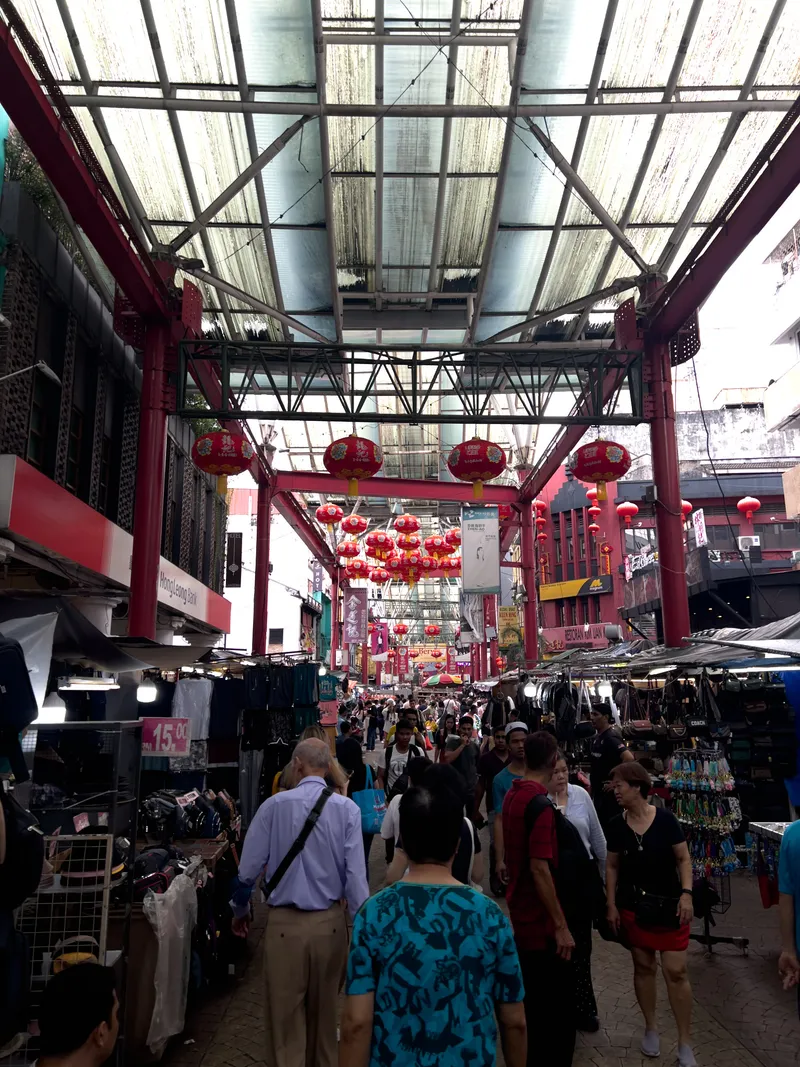 A bustling covered street market in Chinatown, Kuala Lumpur, Malaysia, with crowds of shoppers walking along a narrow lane flanked by vendor stalls on both sides. Red Chinese lanterns hang from metal framework and red steel pillars that support a translucent corrugated roof overhead. Stalls on the left display clothing with price tags showing "15.00," while stalls on the right sell accessories including bags and keychains. A Hong Leong Bank sign is visible on the upper left. The crowd is diverse, including locals and tourists of various ages carrying shopping bags and backpacks. In the background, clusters of red lanterns are grouped together, and traditional Chinese-style architectural elements in red paint frame the market corridor. The atmosphere is vibrant and busy, typical of Petaling Street's famous flea market.