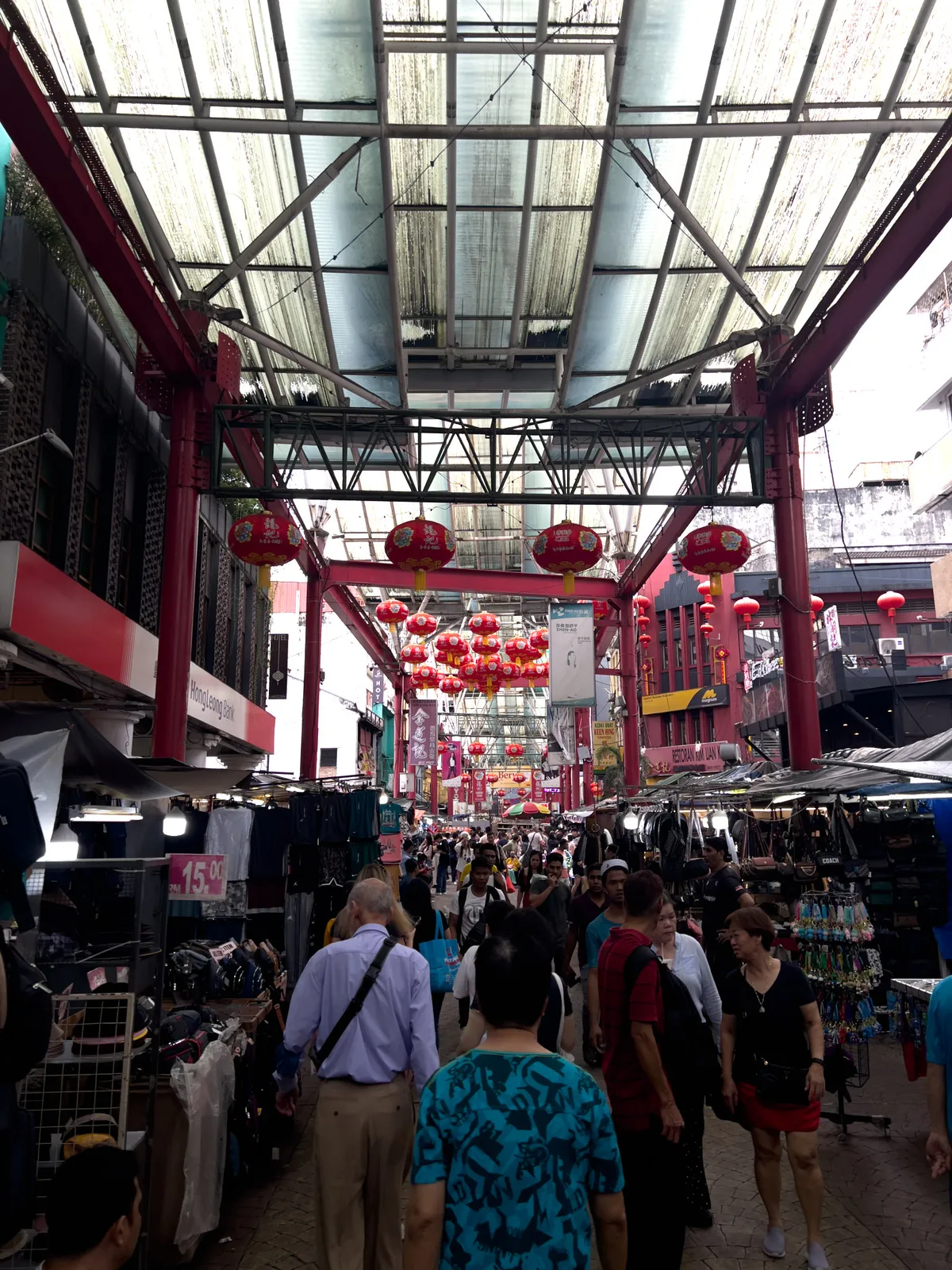 A bustling market scene in Kampung Cendana, Kuala Lumpur, Malaysia. The path is lined with stalls selling clothing, bags and other goods. Red lanterns hang above the market, creating a festive atmosphere. Many people stroll between the stalls, some browsing the wares while others chat. The atmosphere is lively and welcoming, with a blend of modern and traditional elements.
