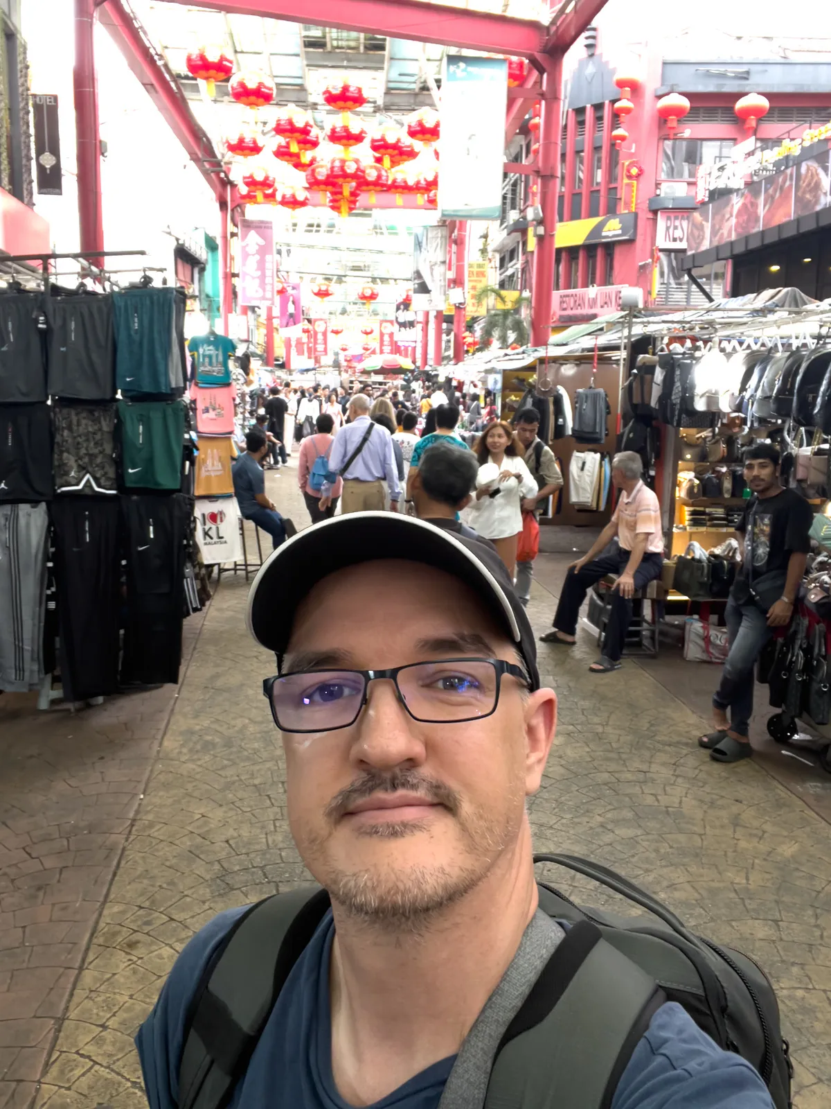 Taken in Kampung Cendana, Kuala Lumpur, this scene shows a bustling market street filled with people. Red lanterns hang above, creating a festive atmosphere. Clothing racks line the left side, while stalls offering various goods complete the picture on the right. The street is paved with patterned brown stone and frequented by passers-by exploring the variety of shops.